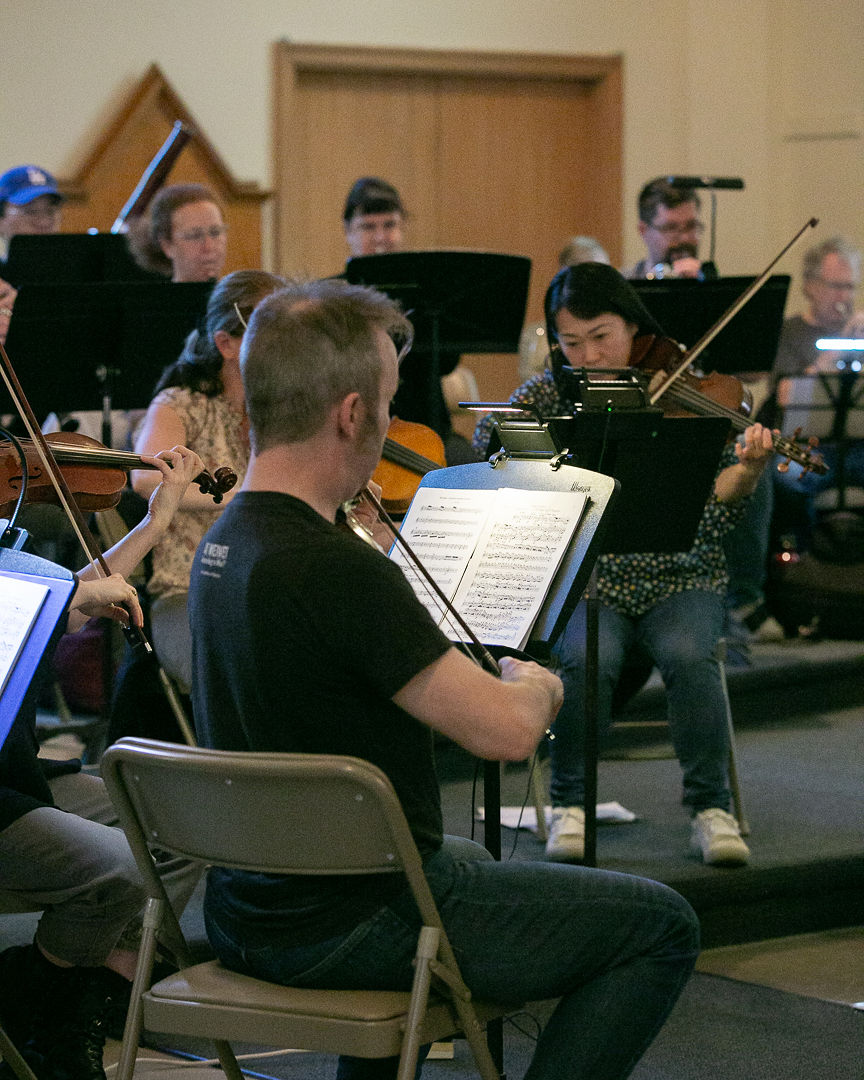 Members of Corktown Community Orchestra play music: violinists and are in the foreground, with various wind and brass players (bassoon, oboe, French horn, trumpet) in the background.